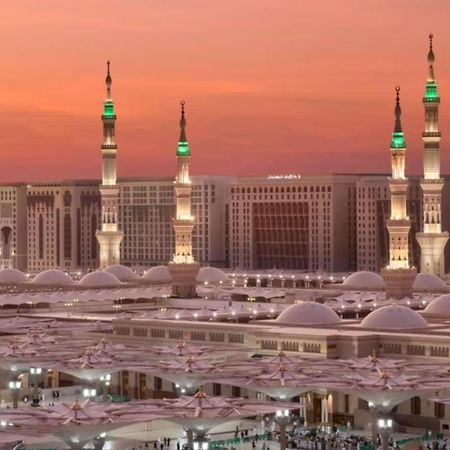 Intercontinental Dar Al Iman Madinah By Ihg Hotel Medina Ngoại thất bức ảnh The photo depicts a scenic view of a mosque complex, likely during sunset, with several minarets illuminated and showcasing green lights. The architecture features domed roofs, and the setting includes a backdrop of buildings, all bathed in a warm, o
