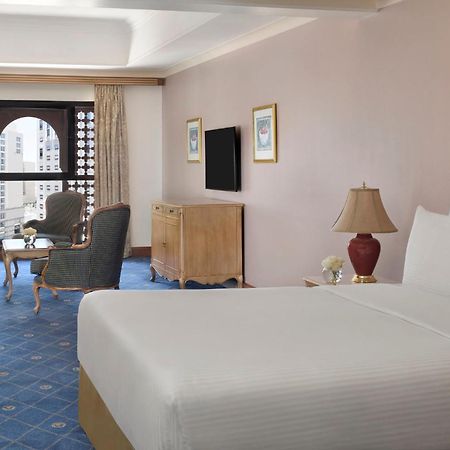 Intercontinental Dar Al Iman Madinah By Ihg Hotel Medina Ngoại thất bức ảnh The photo shows a well-furnished hotel room. It features a large bed with a white duvet, two bedside lamps, and decorative paintings on the walls. There is a comfortable seating area with two chairs and a small table. A wooden dresser is positioned a