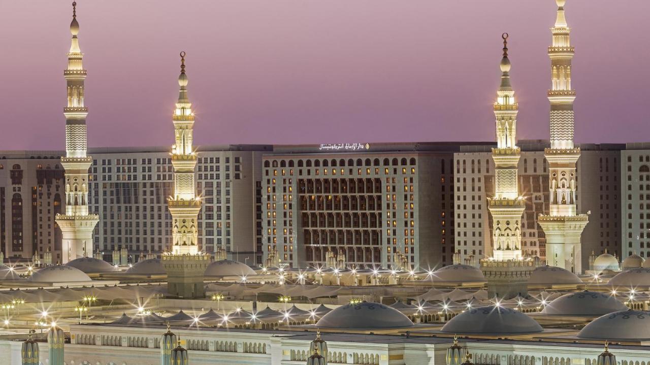Intercontinental Dar Al Iman Madinah By Ihg Hotel Medina Ngoại thất bức ảnh The photo depicts an architectural scene, likely from a mosque complex, featuring prominent minarets that are illuminated. The setting appears to be during twilight, with a soft gradient in the sky. In the background, a large building is visible, pos