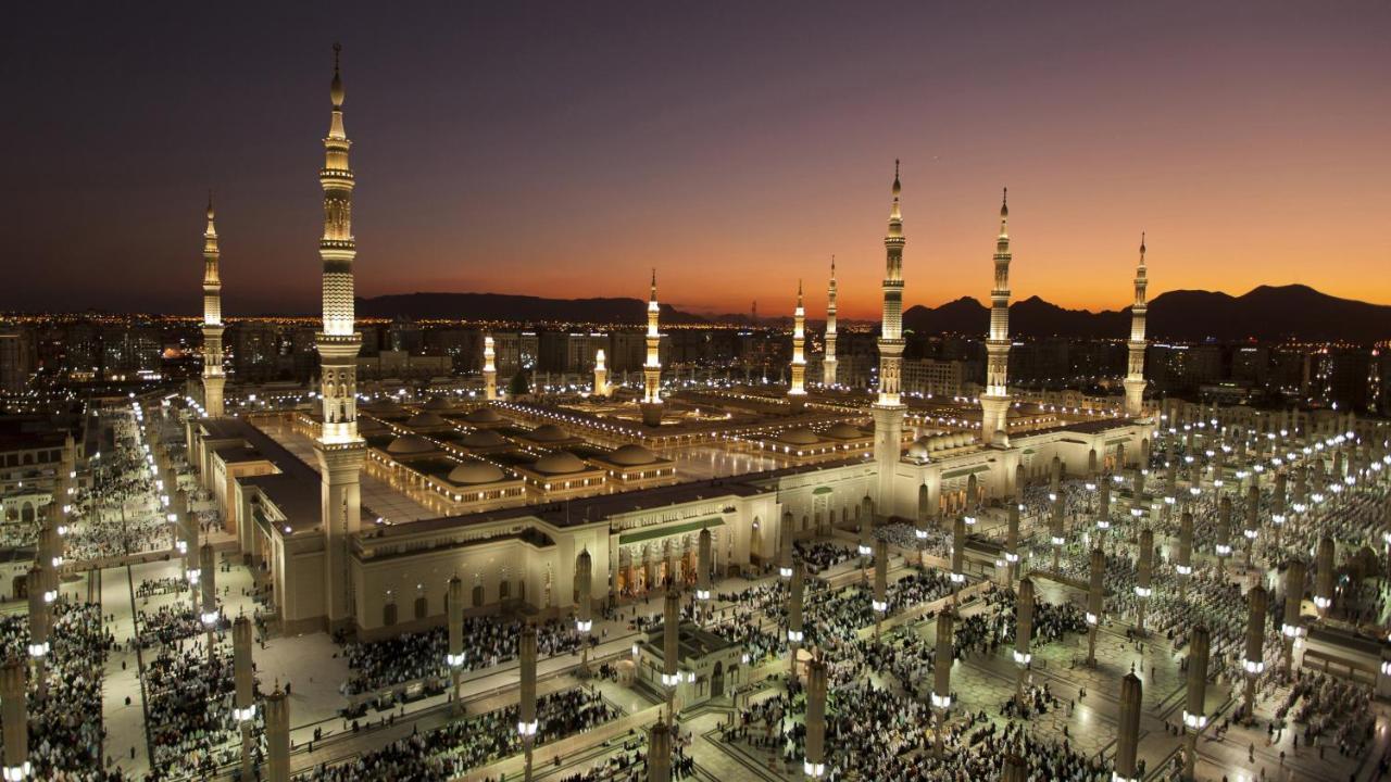Intercontinental Dar Al Iman Madinah By Ihg Hotel Medina Ngoại thất bức ảnh The photo depicts an aerial view of a large mosque complex during sunset. The mosque features multiple tall minarets, which are intricately designed and illuminated, giving the scene a stunning appearance as the sky transitions from daylight to dusk.