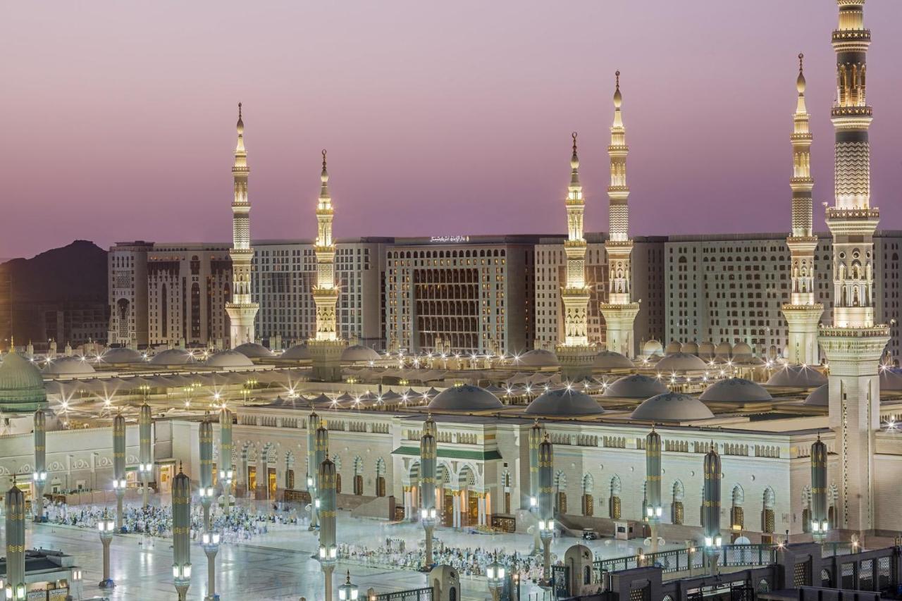 Intercontinental Dar Al Iman Madinah By Ihg Hotel Medina Ngoại thất bức ảnh The photo shows a view of an expansive mosque complex, featuring several tall minarets that are illuminated against a twilight sky. The structure appears grand and is likely adorned with intricate architectural details. There are people gathered in t