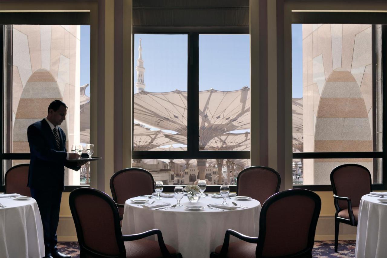 Intercontinental Dar Al Iman Madinah By Ihg Hotel Medina Ngoại thất bức ảnh The photo shows a dining area with elegant decor. A waiter is standing to the side, holding a tray. The tables are neatly set with white tablecloths and fine dining arrangements. Large windows provide a view of an outdoor area featuring architectural