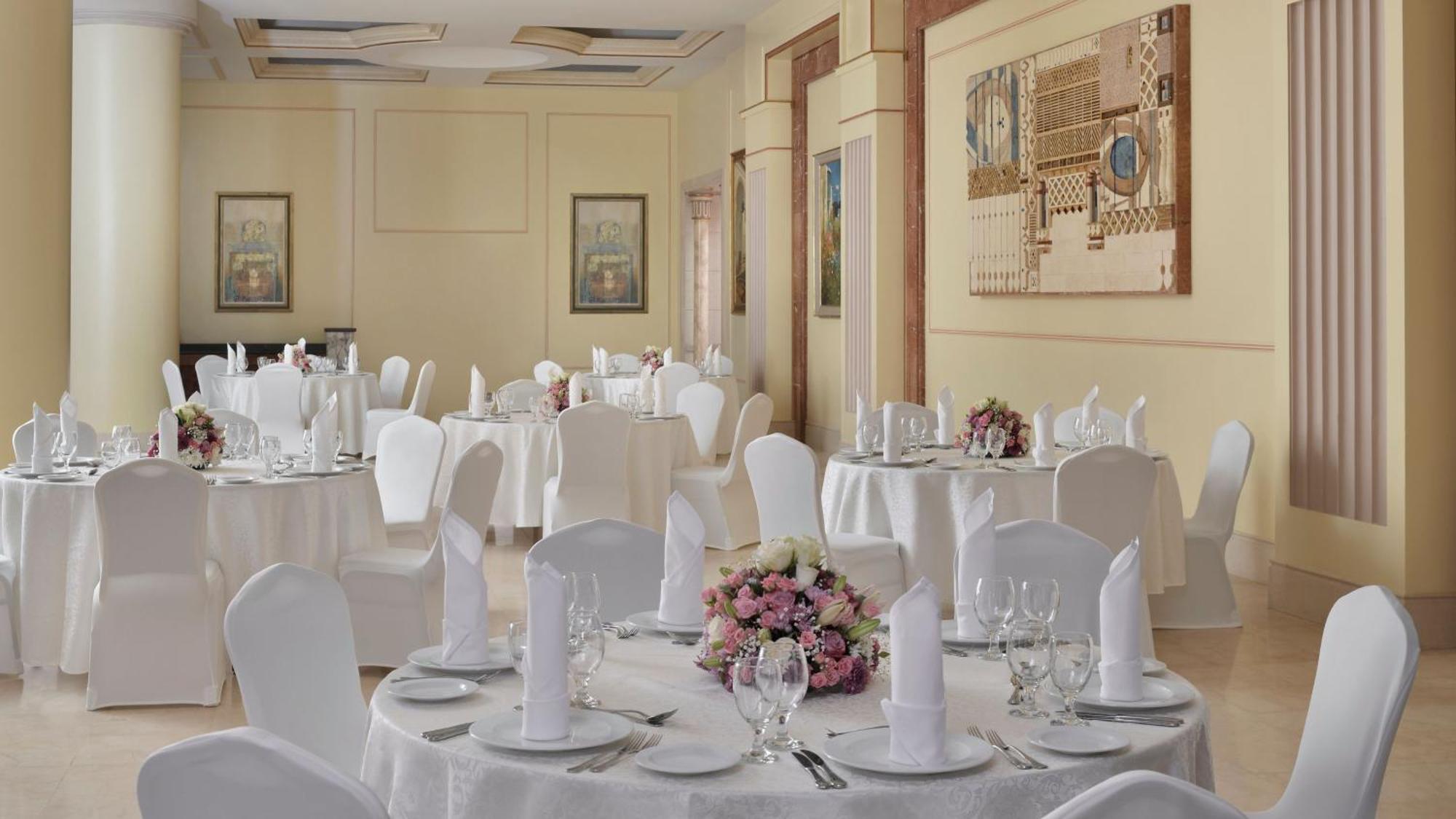 Intercontinental Dar Al Iman Madinah By Ihg Hotel Medina Ngoại thất bức ảnh The photo shows an elegantly arranged dining area, likely set up for a formal event or reception. There are several round tables covered with white tablecloths, each adorned with floral centerpieces featuring pink flowers. The tables are set with pla