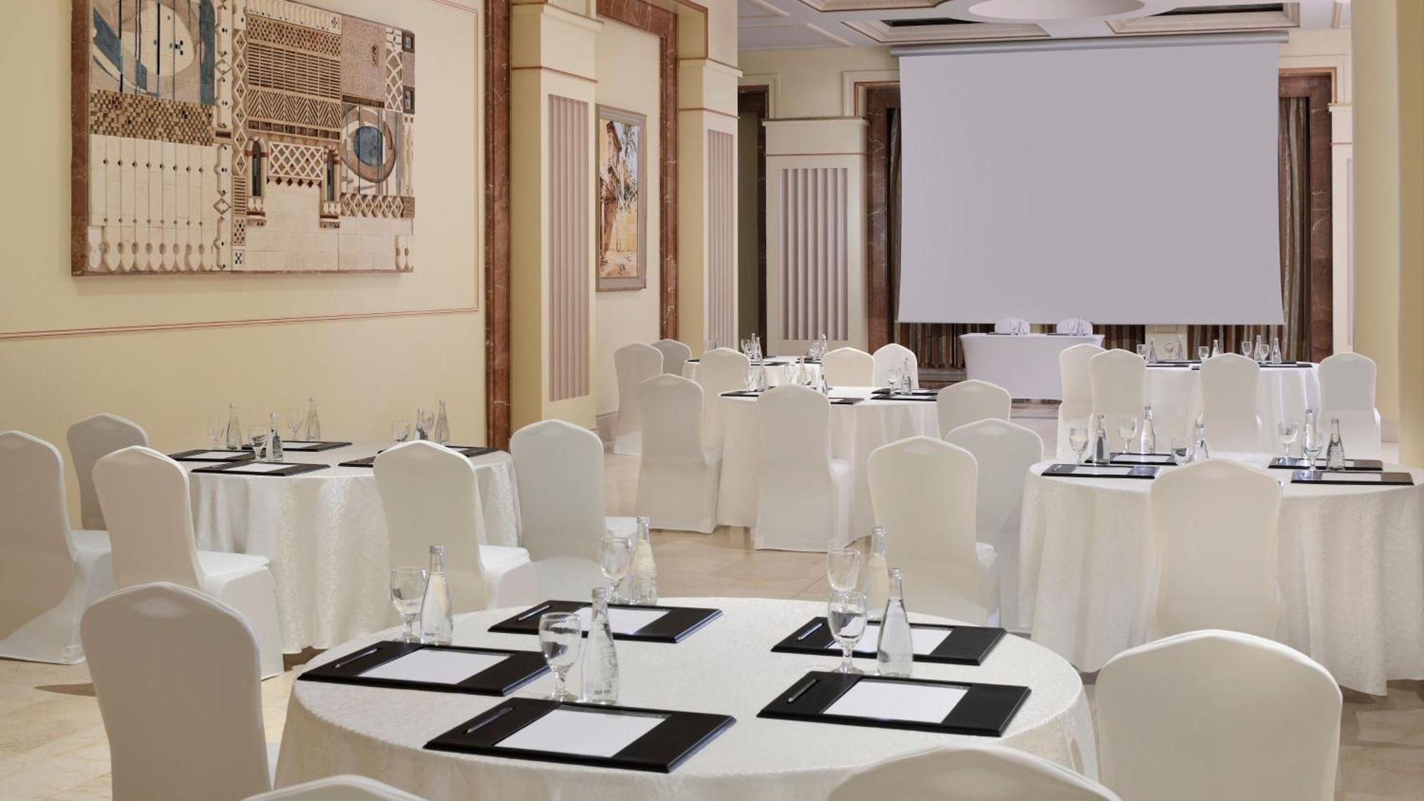 Intercontinental Dar Al Iman Madinah By Ihg Hotel Medina Ngoại thất bức ảnh The photo depicts an elegantly arranged event space. There are several round tables covered with white tablecloths and surrounded by white chairs. Each table is set with a black folder or menu and a glass of water. The walls are adorned with artworks