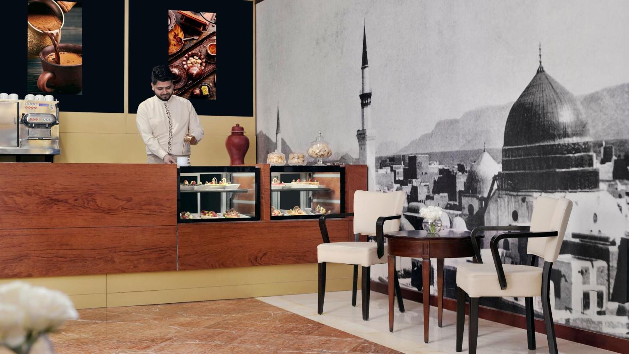 Intercontinental Dar Al Iman Madinah By Ihg Hotel Medina Ngoại thất bức ảnh The photo shows an elegant cafe or dessert shop interior. A man in traditional attire is standing behind a wooden counter, where various pastries and sweets are displayed. The walls feature a large black-and-white mural of a cityscape, possibly depic