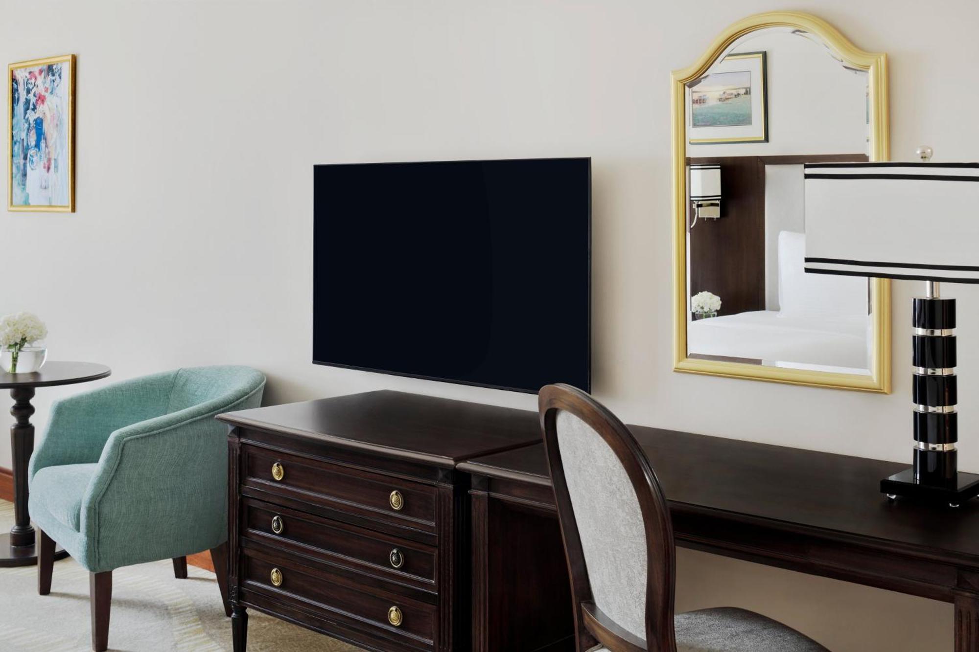 Intercontinental Dar Al Iman Madinah By Ihg Hotel Medina Ngoại thất bức ảnh The photo shows a hotel room interior featuring a mounted flat-screen television on the wall. Below the TV, there is a dark wooden dresser with drawers. To the right, there's a wooden desk and a chair with a light-colored seat. A small table with a l
