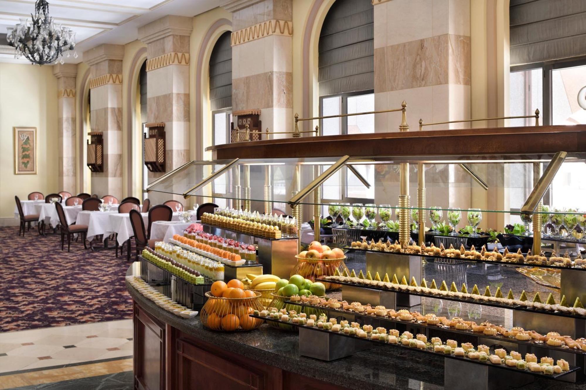 Intercontinental Dar Al Iman Madinah By Ihg Hotel Medina Ngoại thất bức ảnh The photo depicts an elegant dining area featuring a buffet-style display. In the foreground, a beautifully arranged buffet counter showcases an array of desserts, including miniature pastries and colorful fruit. The setting has a sophisticated ambia