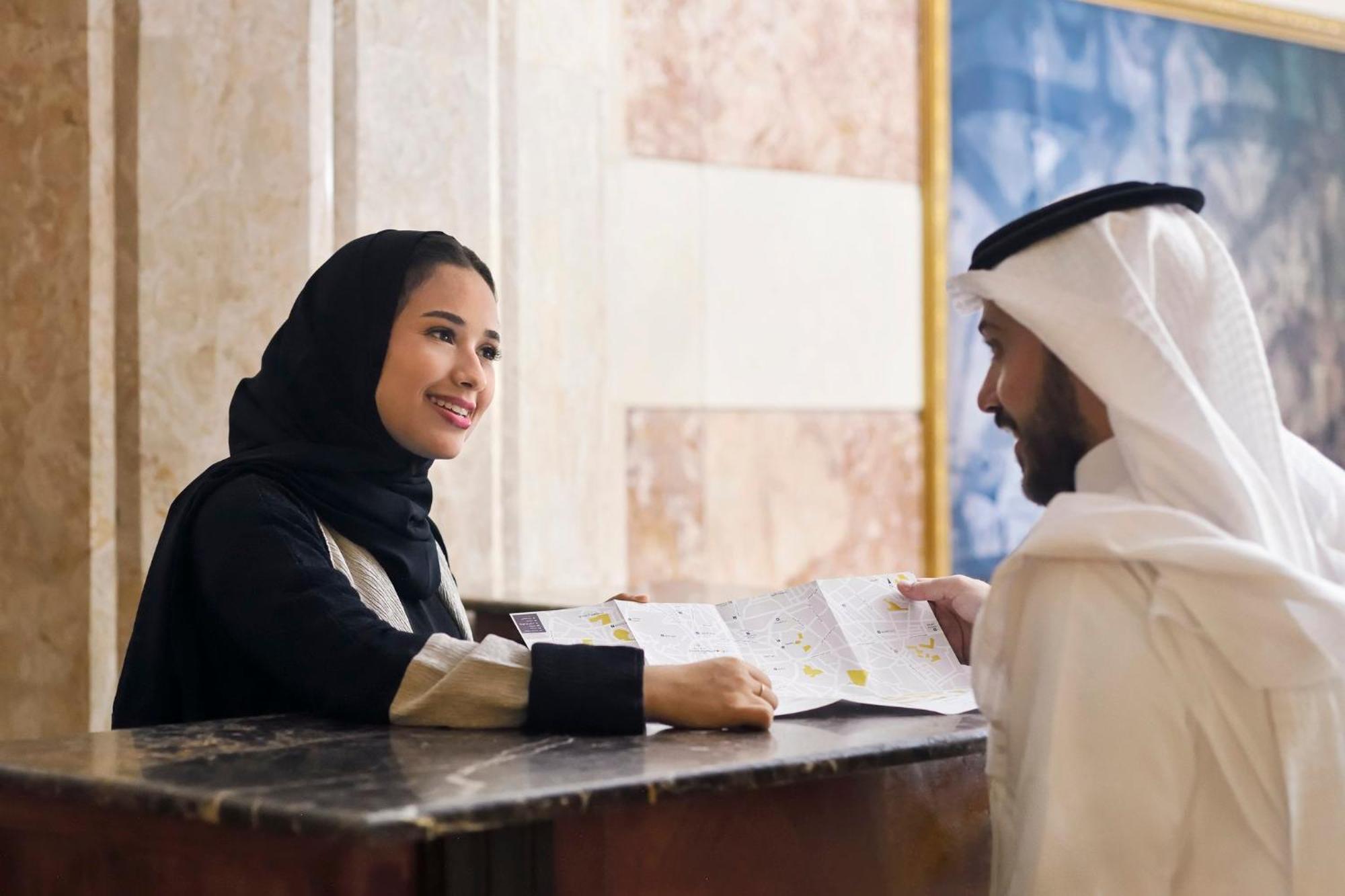 Intercontinental Dar Al Iman Madinah By Ihg Hotel Medina Ngoại thất bức ảnh The photo depicts a woman in a black hijab and a light-colored outfit smiling as she interacts with a man wearing traditional Emirati attire, including a white kandura and ghutrah (headscarf). They are at a reception desk in what appears to be a hote