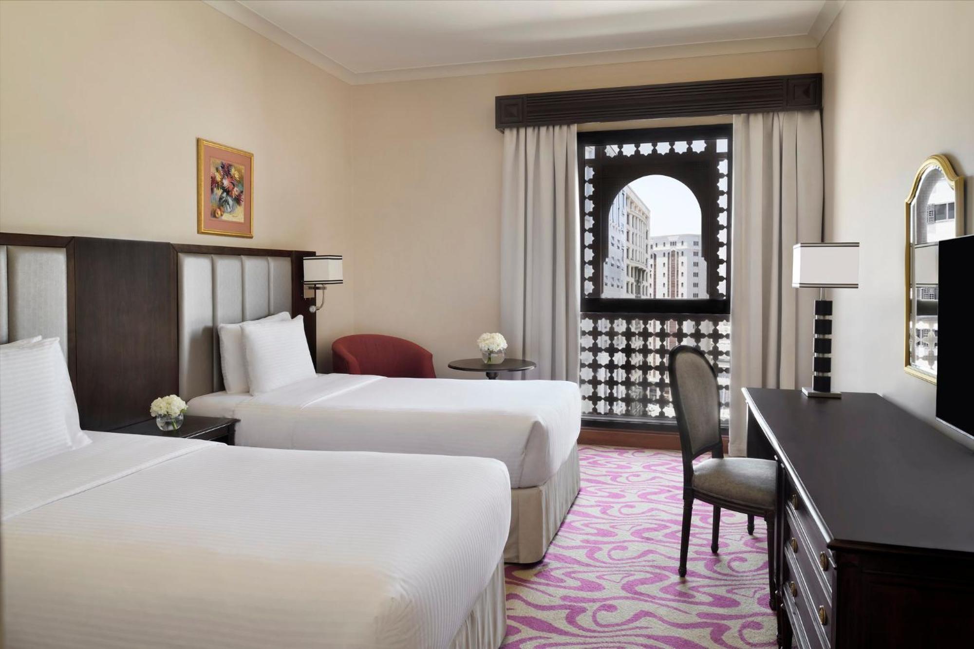 Intercontinental Dar Al Iman Madinah By Ihg Hotel Medina Ngoại thất bức ảnh The photo shows a hotel room featuring two single beds with white bedding. The room has a light, neutral color scheme and is decorated with minimal furnishings. There's a small seating area with a red chair and a round table, as well as a desk with a