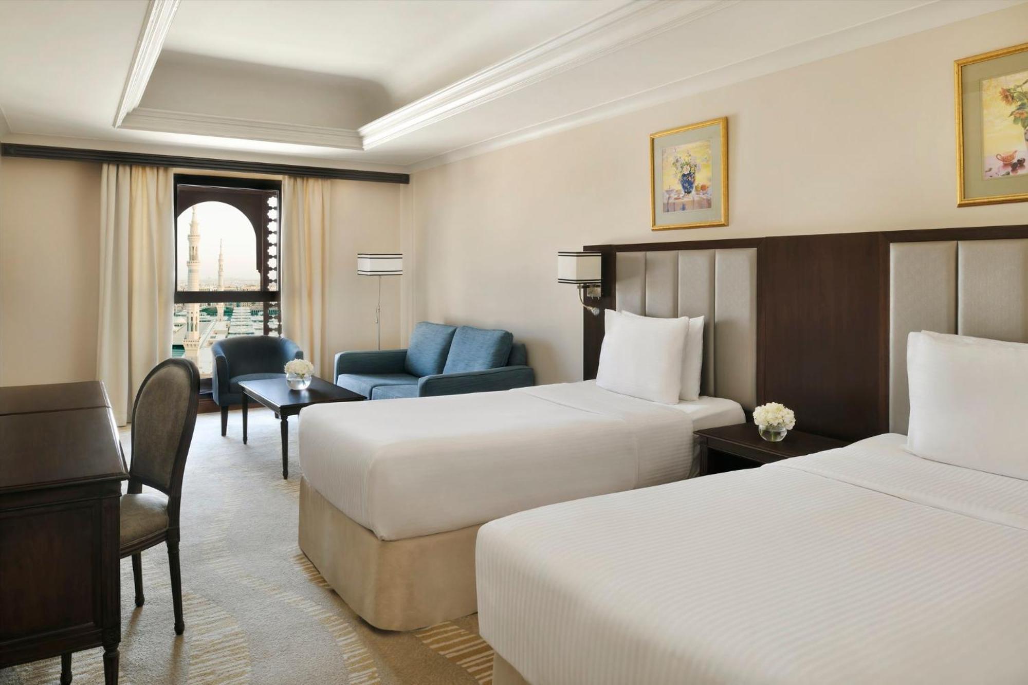 Intercontinental Dar Al Iman Madinah By Ihg Hotel Medina Ngoại thất bức ảnh The photo shows a well-appointed hotel room featuring two twin beds with white bedding. There is a small desk with a chair, a comfortable blue sofa, and a coffee table in the sitting area. The room has a warm, neutral color scheme, accented by two pa