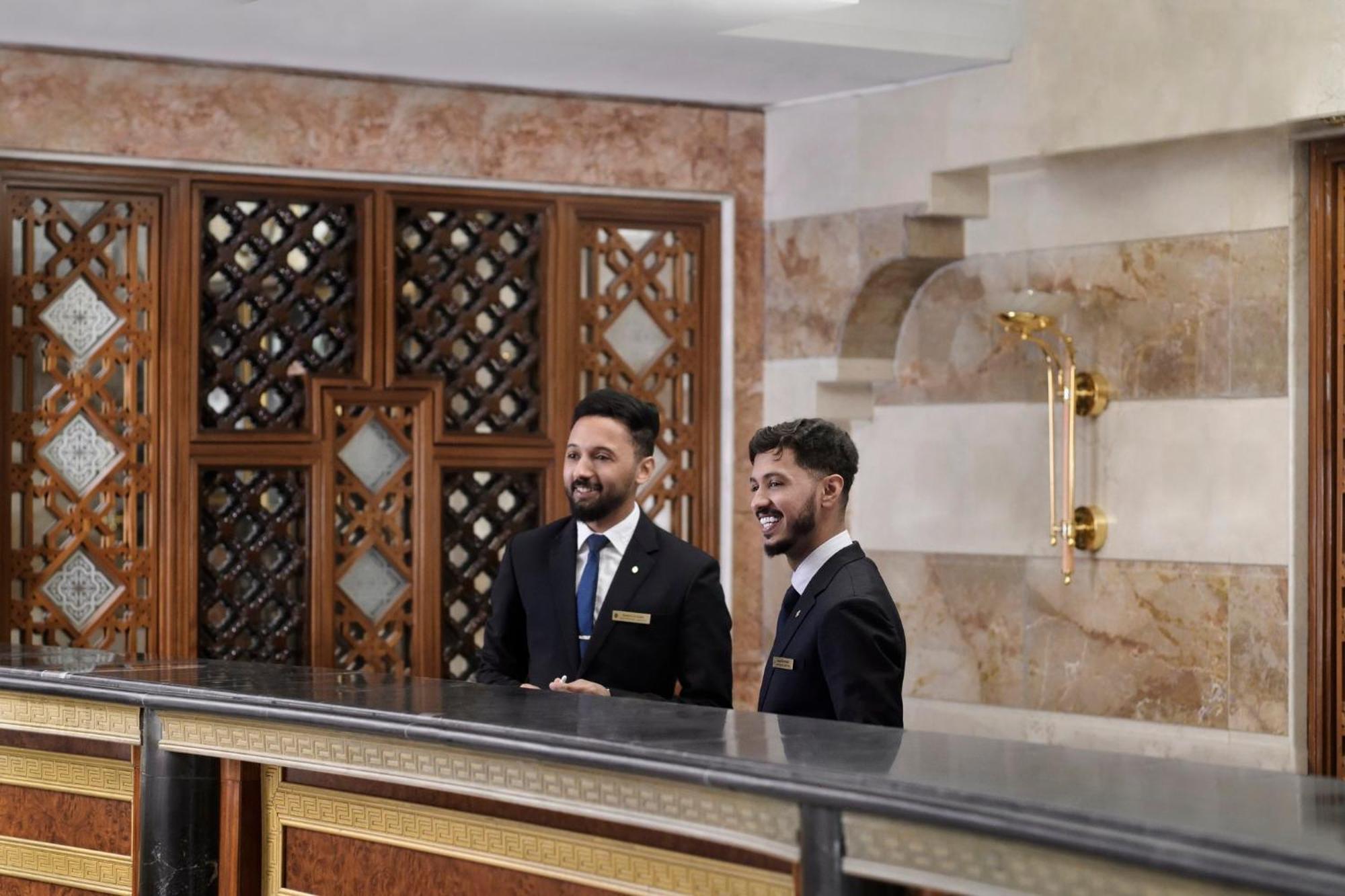 Intercontinental Dar Al Iman Madinah By Ihg Hotel Medina Ngoại thất bức ảnh The photo shows two men standing behind a reception desk in an elegant setting, likely a hotel or similar establishment. They are both dressed in formal attire, wearing suits and ties, and appear to be smiling or engaging in conversation. The backgro