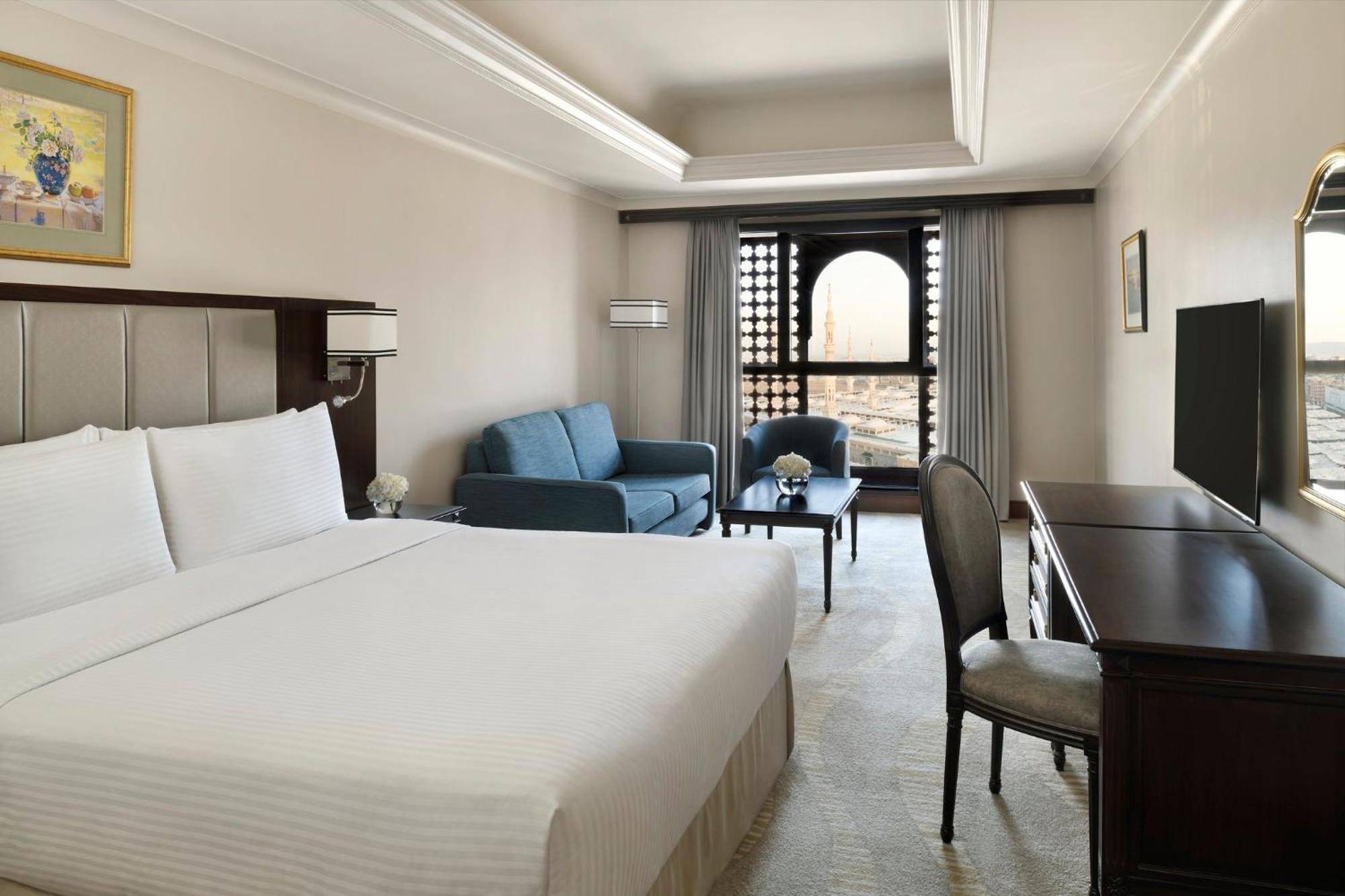 Intercontinental Dar Al Iman Madinah By Ihg Hotel Medina Ngoại thất bức ảnh The photo shows a hotel room designed with a modern and comfortable aesthetic. It features a large bed with white linens, a seating area with a blue sofa, and a small table. There is a wooden desk with a chair and a television opposite the bed. Natur