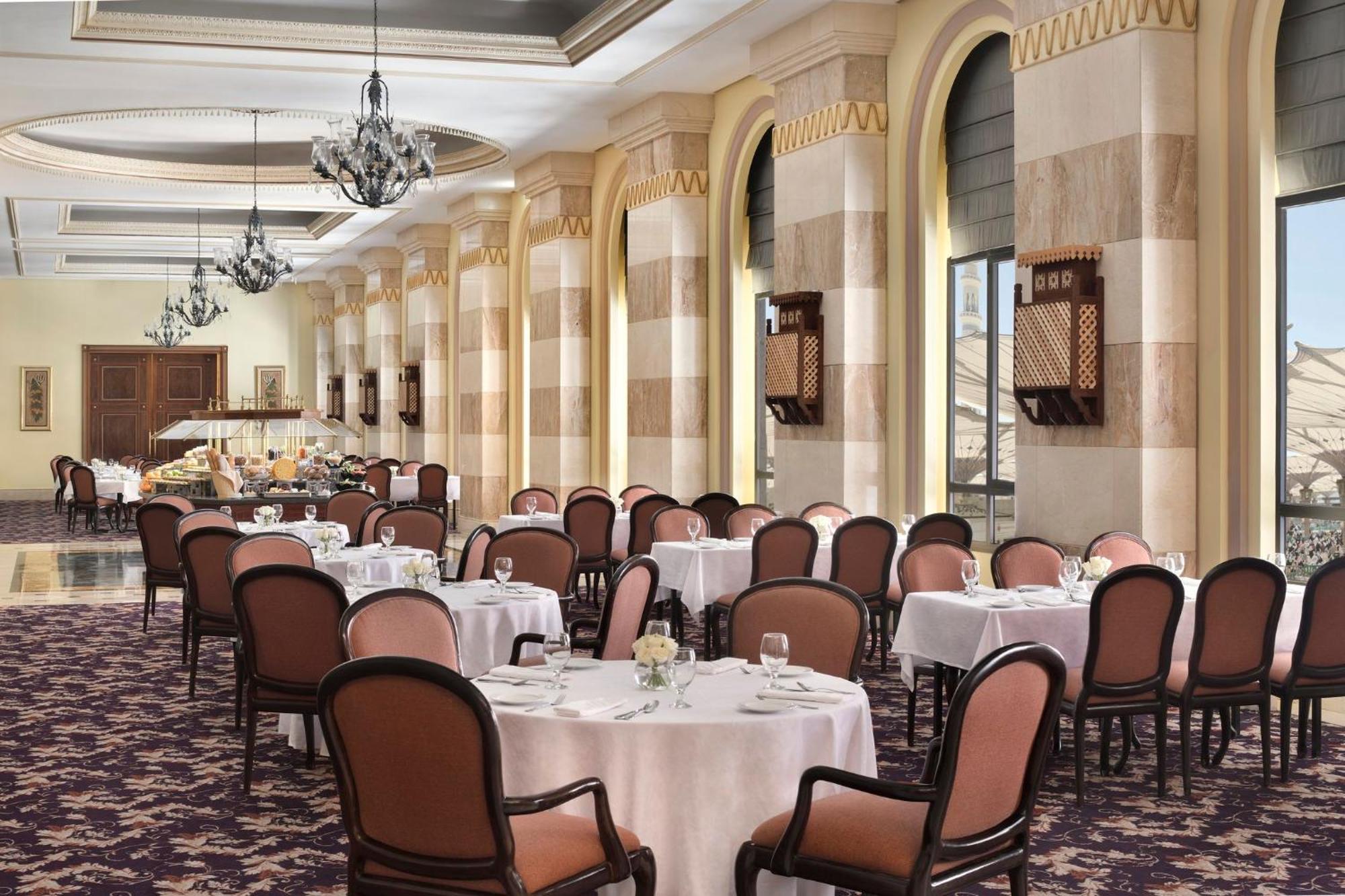 Intercontinental Dar Al Iman Madinah By Ihg Hotel Medina Ngoại thất bức ảnh The photo depicts an elegant dining room setting. It features several round tables covered with white tablecloths, each set with dishes and glassware. The room has a sophisticated ambiance, characterized by high ceilings adorned with chandeliers and 