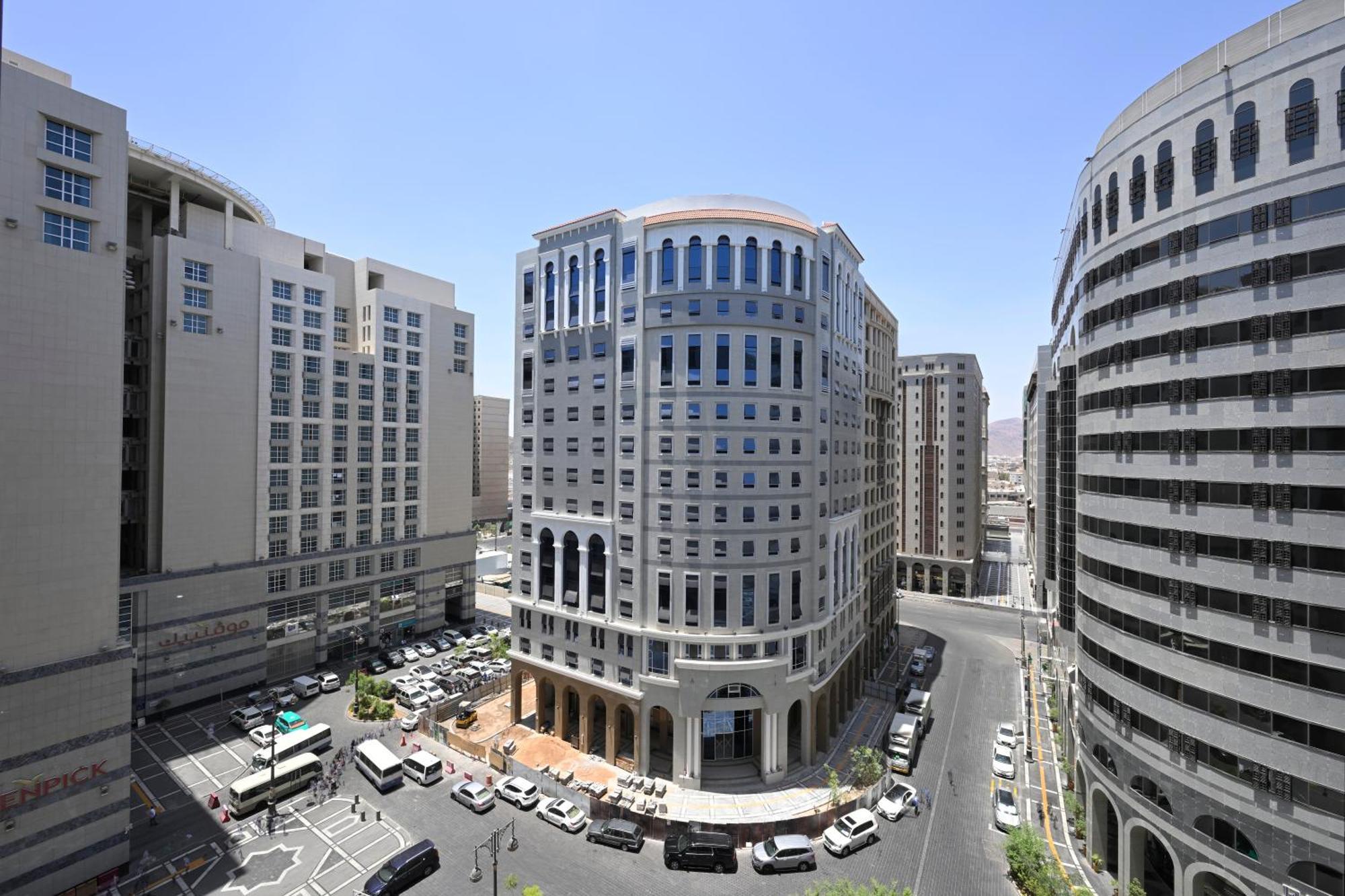 Intercontinental Dar Al Iman Madinah By Ihg Hotel Medina Ngoại thất bức ảnh The photo shows a modern urban scene featuring several buildings, including a prominent rounded building with a glass façade. This building appears to be situated at an intersection, surrounded by other high-rise structures that have distinct archite