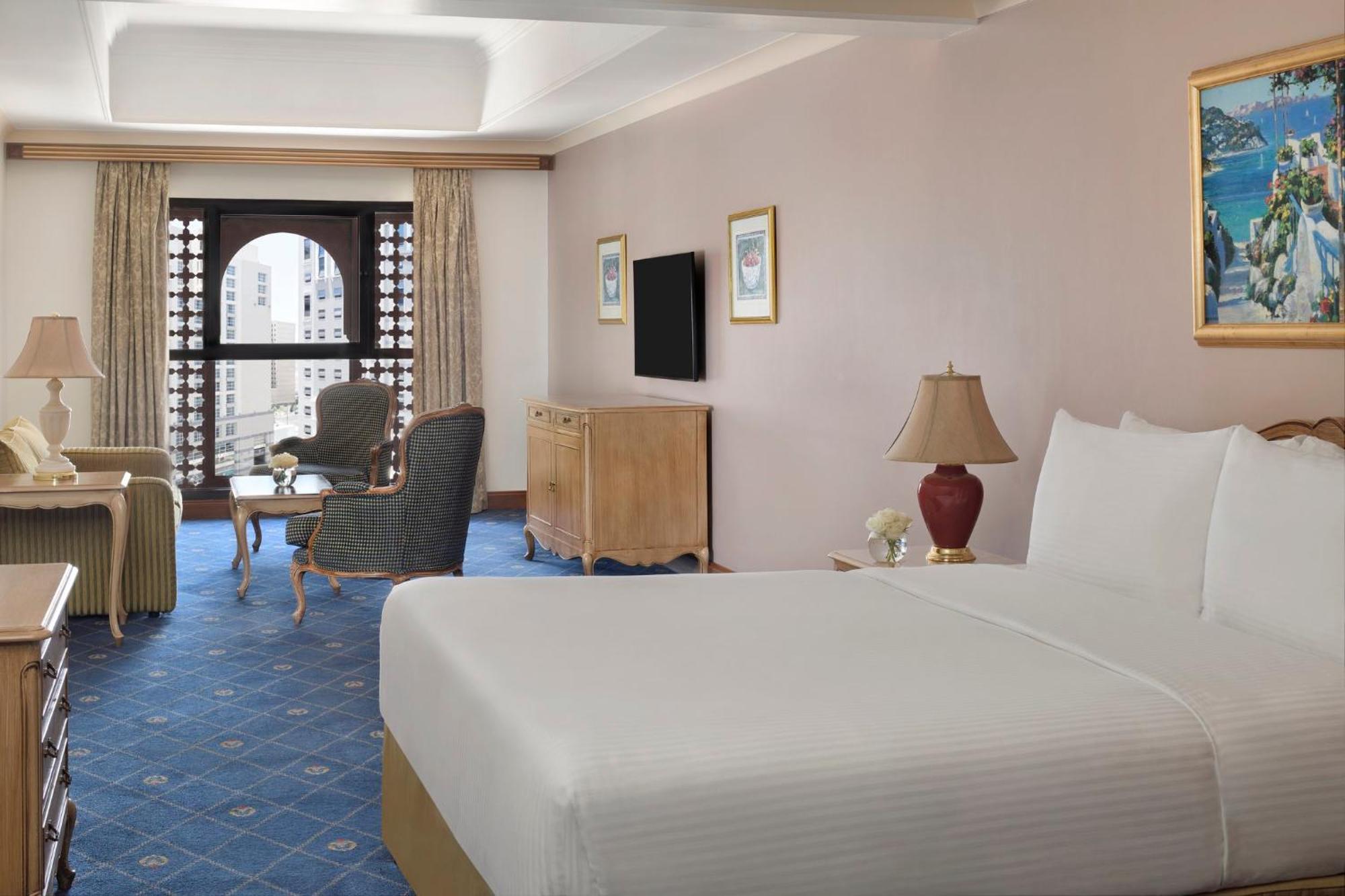 Intercontinental Dar Al Iman Madinah By Ihg Hotel Medina Ngoại thất bức ảnh The photo shows a well-furnished hotel room. It features a large bed with a white duvet, two bedside lamps, and decorative paintings on the walls. There is a comfortable seating area with two chairs and a small table. A wooden dresser is positioned a