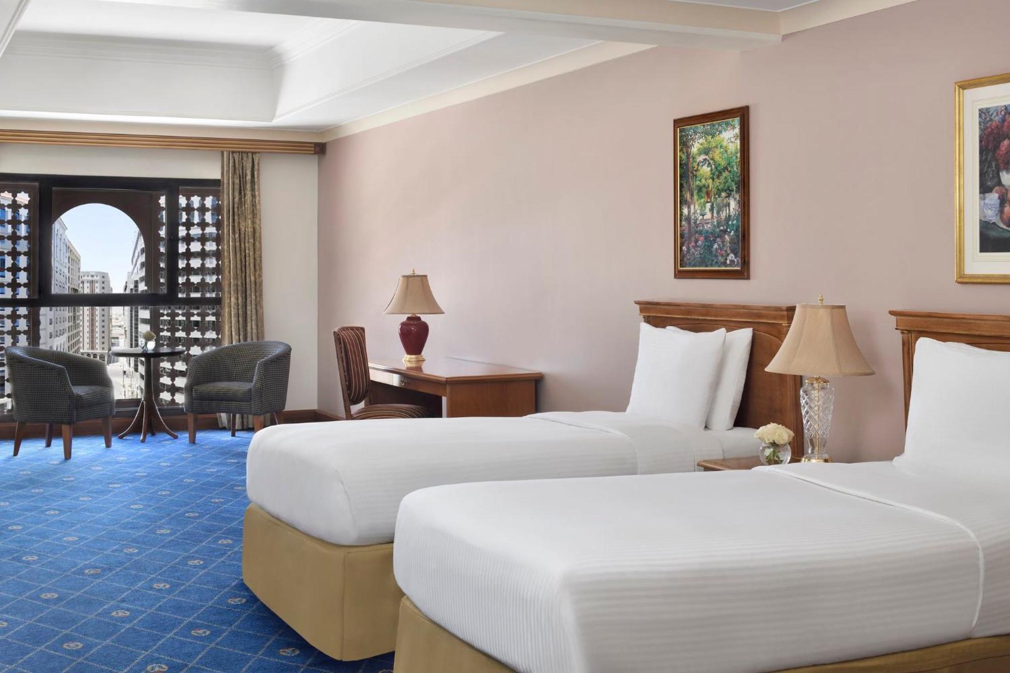 Intercontinental Dar Al Iman Madinah By Ihg Hotel Medina Ngoại thất bức ảnh The photo shows a nicely designed hotel room featuring two twin beds with white linens and beige bases. In the background, there's a wooden desk and a lamp, along with a pair of comfortable armchairs positioned near a large window. The window showcas
