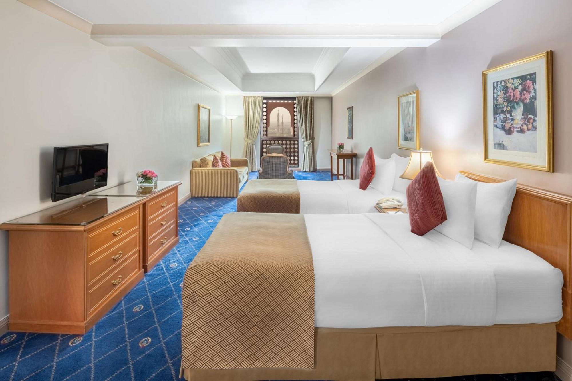 Intercontinental Dar Al Iman Madinah By Ihg Hotel Medina Ngoại thất bức ảnh The photo shows a hotel room featuring two beds with white linens and decorative pillows. There is a wooden dresser with a television on it. The flooring has a blue carpet with a pattern, and the walls are painted in a light color. The room is well-l