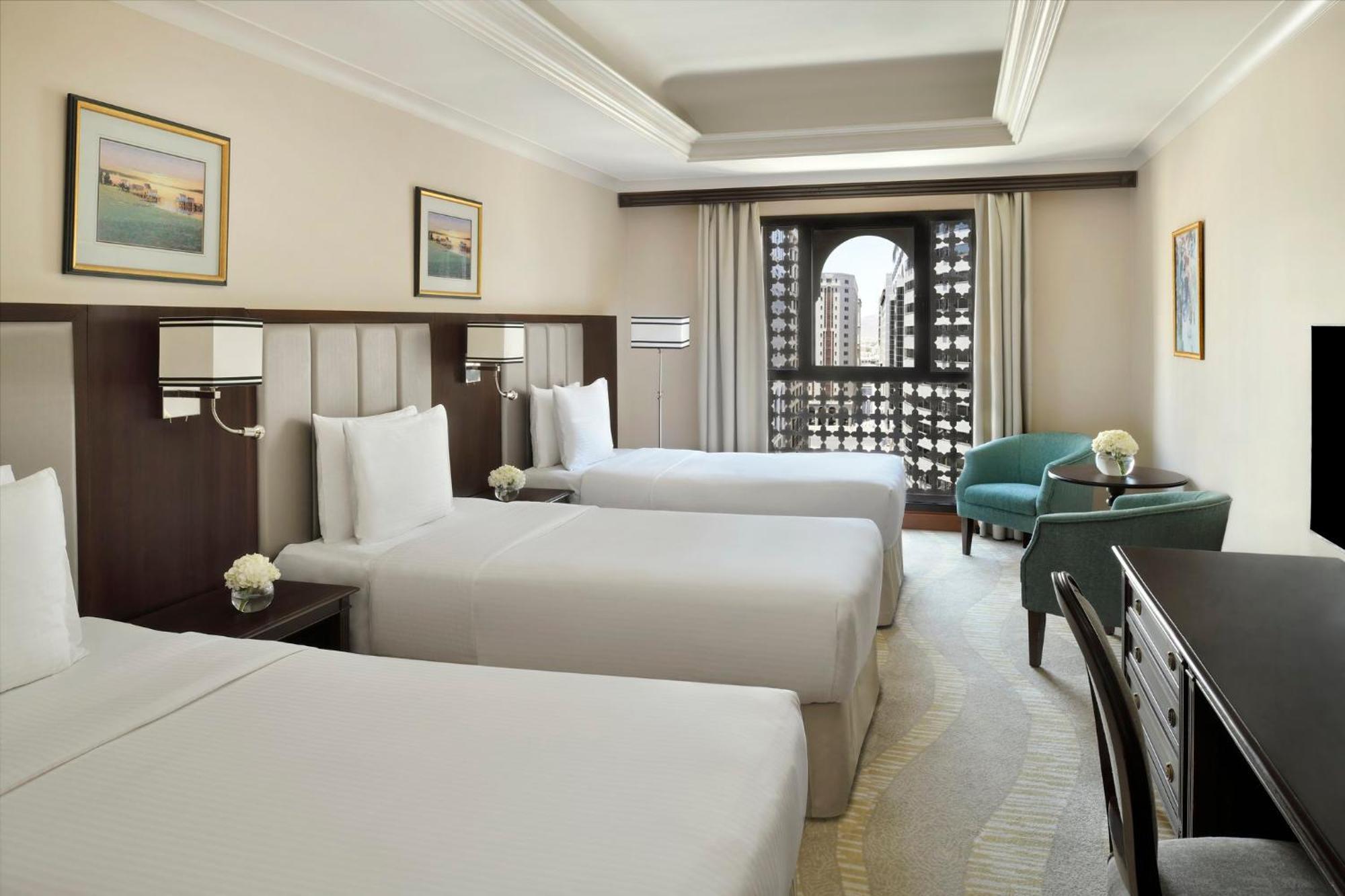 Intercontinental Dar Al Iman Madinah By Ihg Hotel Medina Ngoại thất bức ảnh The photo shows a hotel room with three single beds, dressed in white linens. There are two paintings on the walls, depicting serene landscapes. A large window allows natural light to enter, and it is adorned with elegant curtains. A smaller seating 