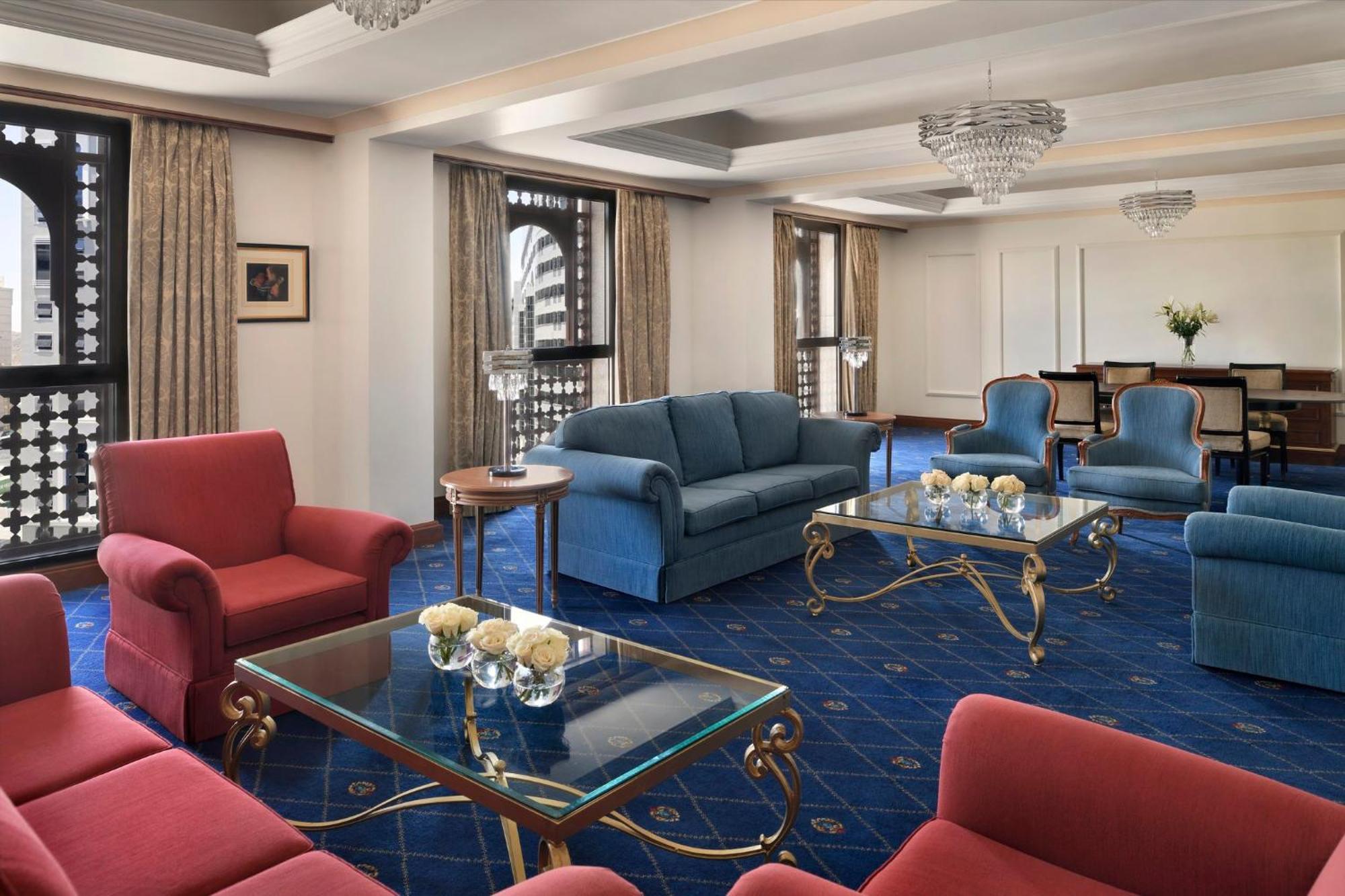 Intercontinental Dar Al Iman Madinah By Ihg Hotel Medina Ngoại thất bức ảnh The photo shows a luxurious living space or lounge area. It features a combination of furniture, including a blue sofa, several red armchairs, and glass-top coffee tables with gold accents. The flooring is carpeted in a deep blue, and the walls are a