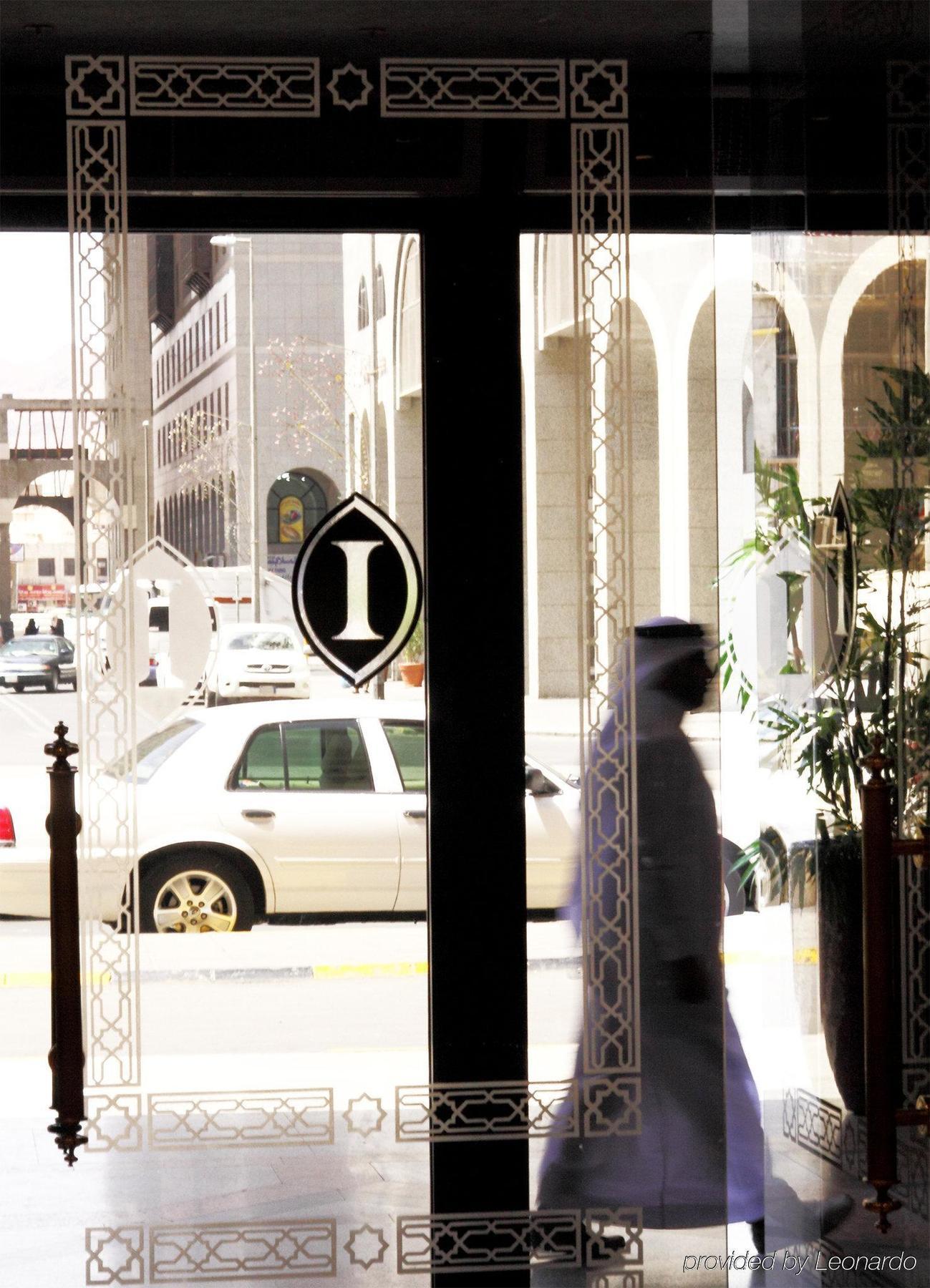 Intercontinental Dar Al Iman Madinah By Ihg Hotel Medina Nội địa bức ảnh The photo shows a view through a glass door or window, featuring an elegant design with a prominent "I" symbol displayed. Through the glass, there are some parked vehicles visible, including a white car. In the foreground, a shadowy figure of a perso