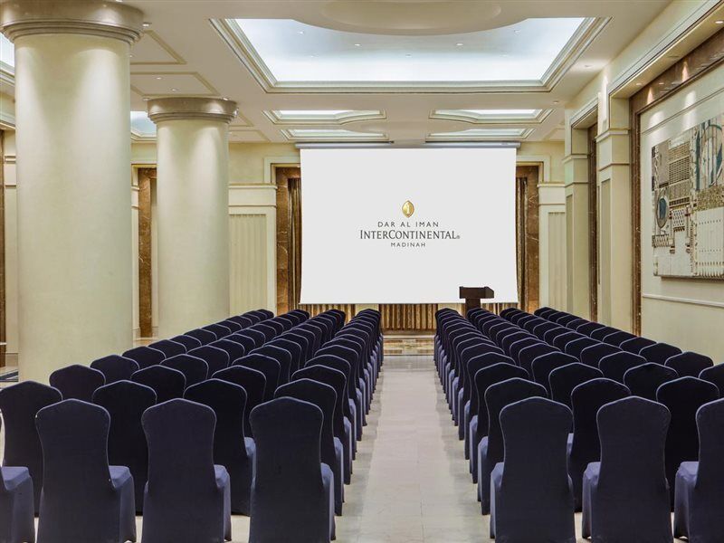 Intercontinental Dar Al Iman Madinah By Ihg Hotel Medina Ngoại thất bức ảnh The photo shows a conference or meeting room set up. There are rows of dark blue chairs arranged facing a large screen at the front of the room. The screen displays the logo of the Intercontinental hotel, indicating that this space is likely intended