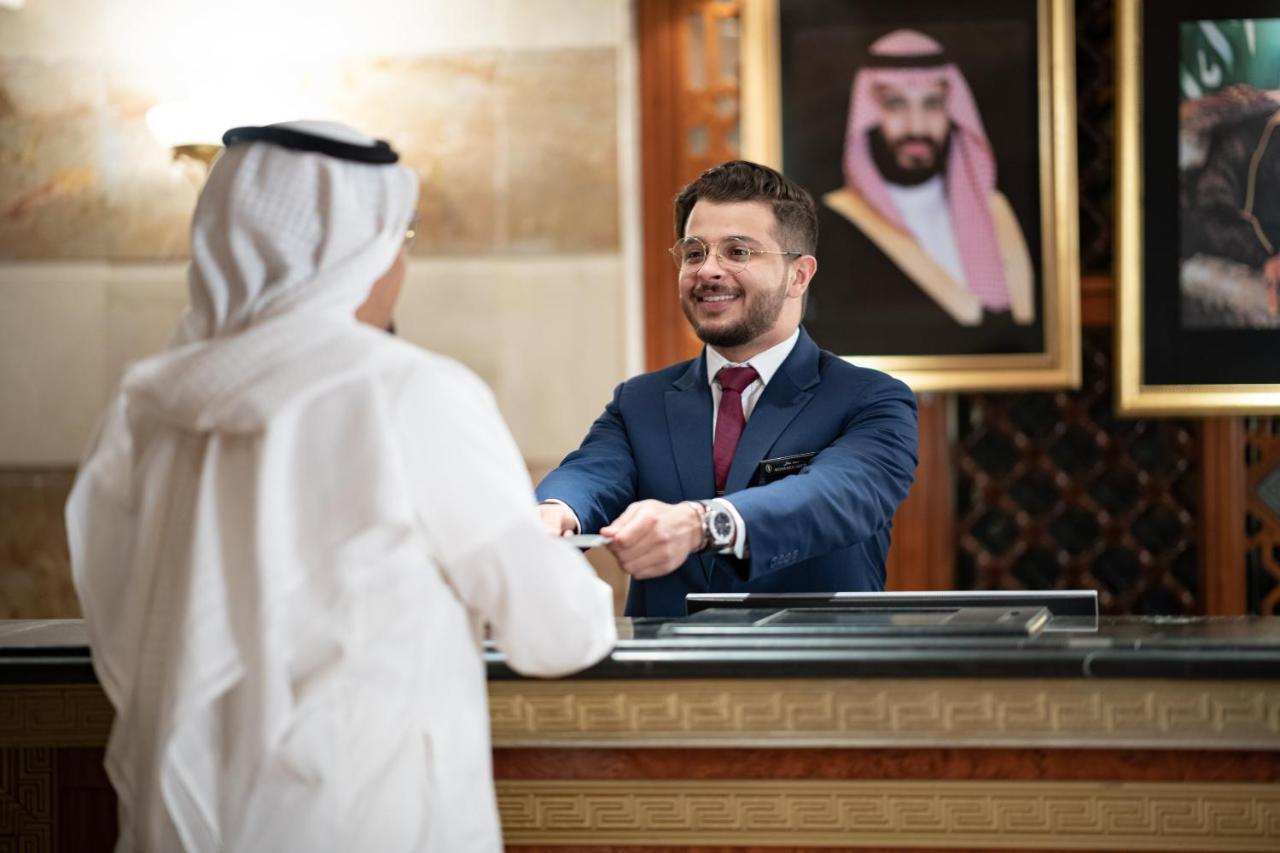 Intercontinental Dar Al Iman Madinah By Ihg Hotel Medina Ngoại thất bức ảnh The photo shows a hotel reception area. A male receptionist, dressed in a navy suit with a tie, is smiling and handing an item to a guest who is wearing traditional white attire, possibly a kandura or thobe. The background features elegantly decorate