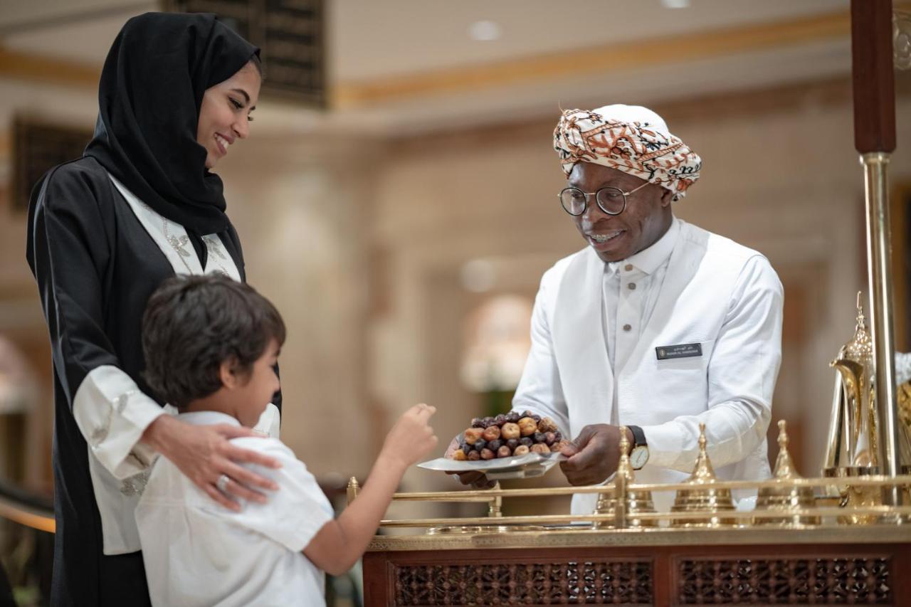 Intercontinental Dar Al Iman Madinah By Ihg Hotel Medina Ngoại thất bức ảnh The photo depicts a warm interaction between three people. A woman, dressed in a traditional black attire and a headscarf, stands beside a young boy who is reaching out towards a plate of dates held by a smiling man in a white traditional outfit with
